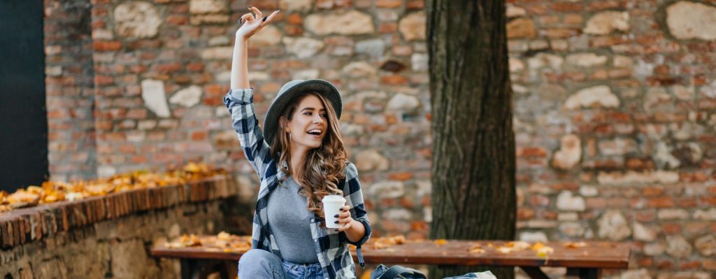 A woman with a hat holding a cup of coffee and waving in a park in fall